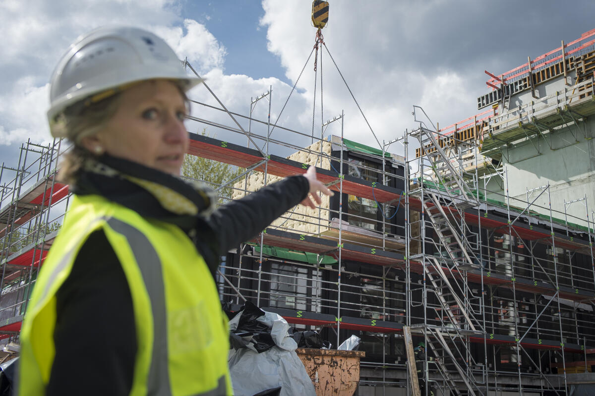  Begehung der Baustelle Luisenblock West Modulbau - Petra Wesseler, Präsidentin für Bauwesen und Raumordnung, zeigt auf ein Holzmodul, was gerade mit einem Kran eingehoben wird.