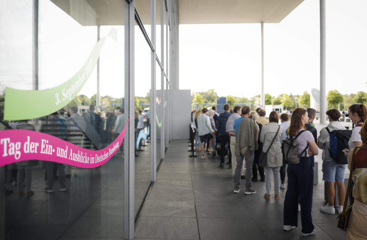  Bunte Banner an der Glasfassade des Paul-Löbe-Hauses weisen auf den Tag der Ein- und Ausblicke des Deutschen Bundestages am 3. September 2023 hin. Daneben warten Besucherinnen und Besucher in einer Schlange vor dem Eingang auf Einlass.