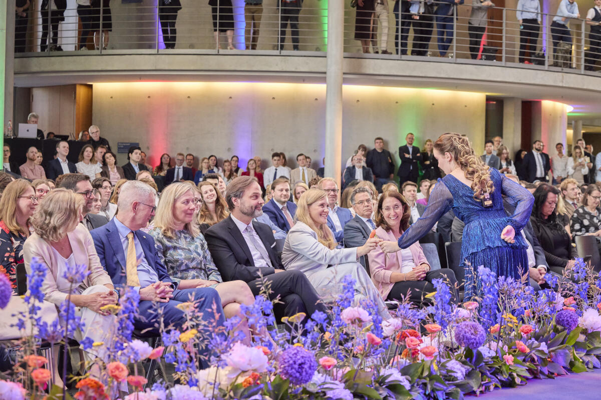 Ortleb, Josephine; Radomski, Kerstin; Göttke, Paul; Özoğuz, Aydan Empfang des Internationalen Parlaments-Stipendiums (IPS) in der Halle des Paul-Löbe-Hauses - Hier bekommt Bundestagsvizepräsidentin Josephine Ortleb (1. Reihe 5vl) im Rahmen des kreativen Bühnenprogramms der Stipendiatinnen und Stipendiaten des IPS global 2026 während der Aufführung der Arie "Mein Herr Marquis" (Lachlied) aus Die Fledermaus von Johann Strauss eine Blume von der Sängerin überreicht.