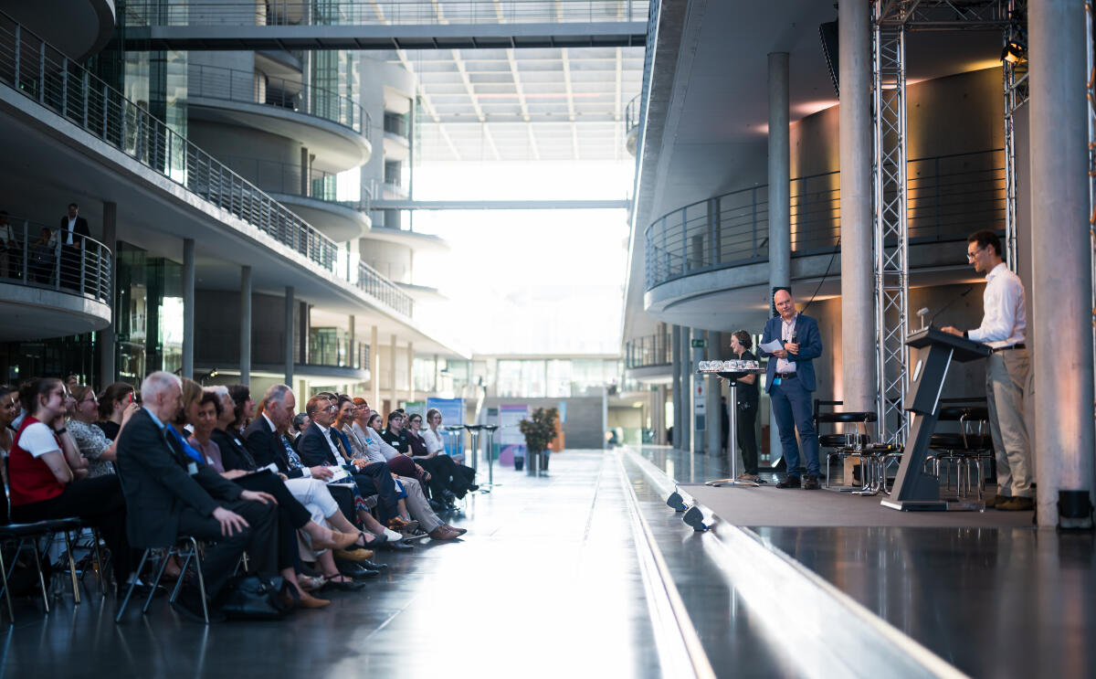  Eröffnung der Ausstellung "Poster im Bundestag: Wissenschaftsfreiheit - die Freiheit zu forschen" in der Halle des Paul-Löbe-Hauses: Dr. Jan-Martin Wiarda (2vr), Journalist und Moderator, moderiert das Bühnenprogramm. Hier mit Karim Walkusch (r), beteiligter Studierender der Humboldt-Universität zu Berlin, beim Elevator Pitch während des Bühnenprogrammes und mit Blick auf das Publikum.
